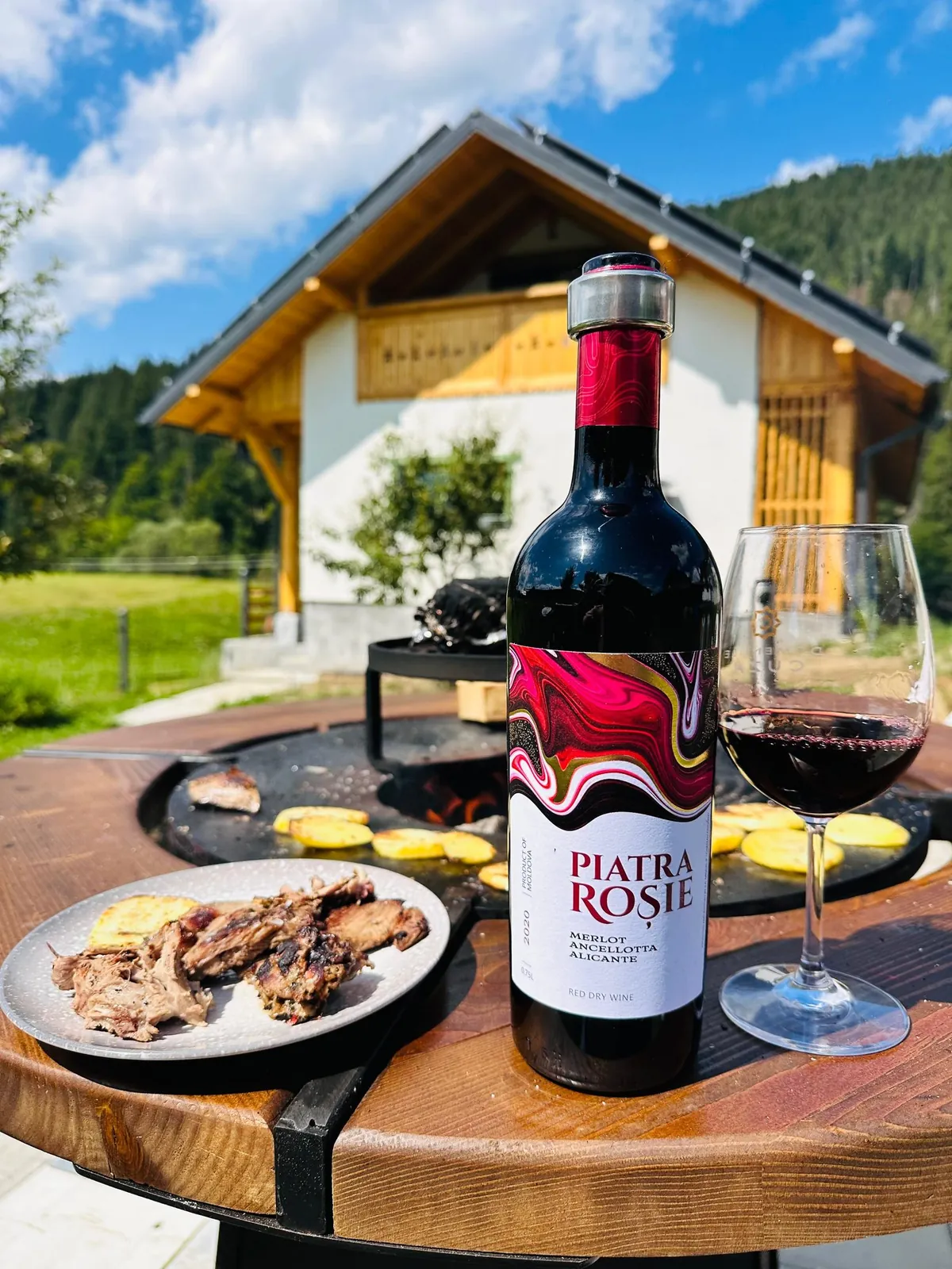 Grătarul rotund din foișor, cu o sticlă de vin și casa în spate