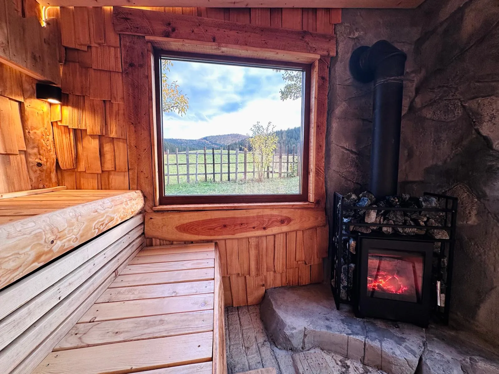 Sauna în amenajare: banca de lemn, soba pe lemne și geamul mare spre pajiște