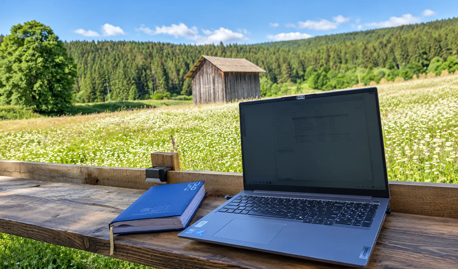 Laptop deschis și o agendă pe o masă de lemn, în fața unei pajiști cu flori, cu o colibă de lemn și pădure de brad în spate